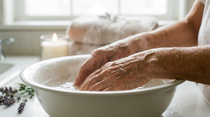 Nahaufnahme der Hände einer älteren Person, die sanft in warmem Wasser in einer weißen Keramikschüssel baden. Entspannte Stimmung, weiches Licht, helles Badezimmer-Setting.