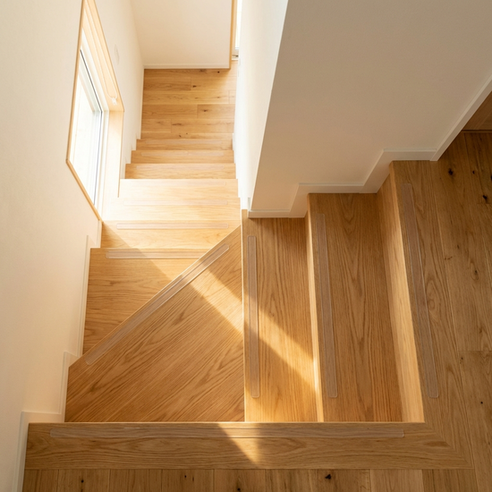 Eine saubere Holztreppe von oben fotografiert, die völlig frei von Stolperfallen ist. Transparente Antirutsch-Streifen sind dezent auf den Stufen erkennbar, warmes Sonnenlicht erhellt den aufgeräumten Raum.