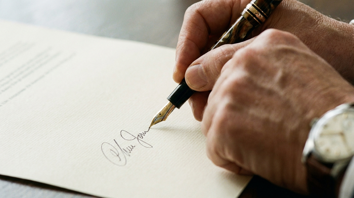 Eine Nahaufnahme einer Hand, die mit einem edlen Füllfederhalter eine Unterschrift auf ein formelles Papier setzt. Klarer Fokus auf den Stift, ohne lesbaren Text.