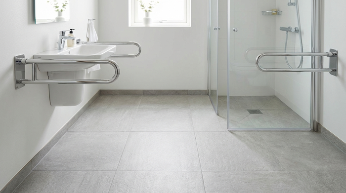 Ein helles, aufgeräumtes Badezimmer mit rutschfesten Bodenfliesen und fest installierten, verchromten Haltegriffen neben dem Waschbecken und der Dusche. Keine Stolperfallen auf dem Boden.