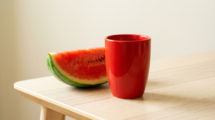 Ein leuchtend roter Trinkbecher steht gut sichtbar auf einem hellen Holztisch neben einem frischen Stück Wassermelone. Klare Kontraste, keine ablenkenden Muster im Hintergrund.