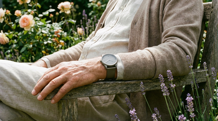 Eine Nahaufnahme einer modernen, unauffälligen Smartwatch am Handgelenk eines älteren Herrn, der entspannt im Garten sitzt. Die Uhr sieht elegant aus und fügt sich natürlich in die Kleidung ein.