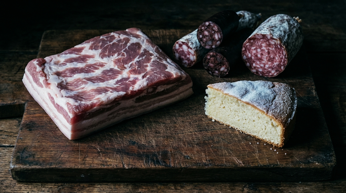 Eine dunklere Szene auf einem hölzernen Schneidebrett: rohes Schweinefleisch, fettige Wurstwaren und ein Stück heller, zuckerreicher Kuchen. Die Beleuchtung ist kühl und wirft harte Schatten.