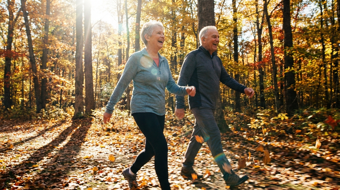 Ein aktives Seniorenpaar spaziert lachend durch einen herbstlichen Wald. Beide tragen sportliche, unauffällige Hörgeräte. Sonnenlicht fällt durch die Blätter. Fokus auf Bewegung und Vitalität.