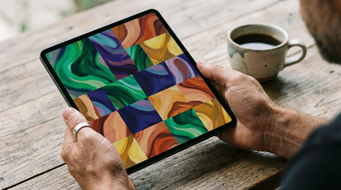 Nahaufnahme von Händen, die ein modernes Tablet halten. Auf dem Bildschirm sind farbige, abstrakte Kacheln zu sehen. Im Hintergrund eine unscharfe Kaffeetasse auf einem Holztisch.