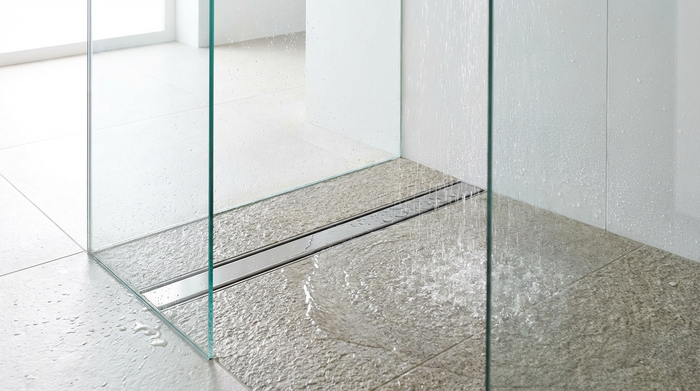 Nahaufnahme einer modernen, bodengleichen Dusche mit eleganter Glaswand. Das Wasser läuft sanft über rutschfeste, strukturierte Fliesen in einen flachen Edelstahl-Abfluss. Helle und saubere Umgebung ohne Text.