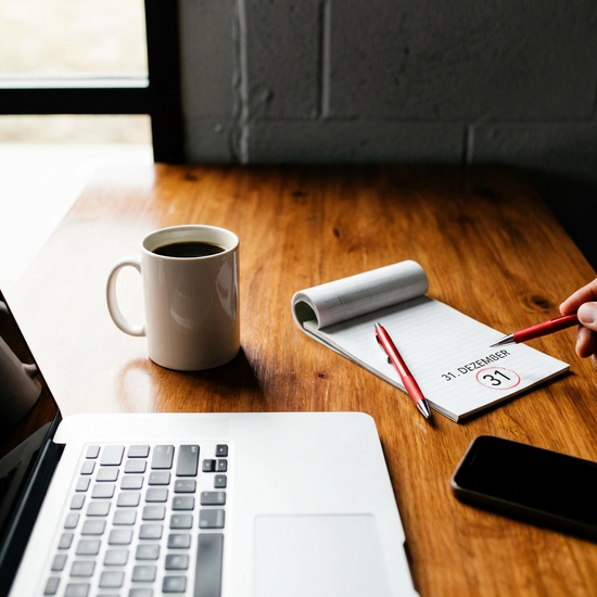 Ein moderner Wandkalender, bei dem ein roter Stift auf den 31. Dezember zeigt. Im Hintergrund unscharf ein aufgeräumter Schreibtisch mit einer Kaffeetasse. Fokus auf das markierte Jahresende.