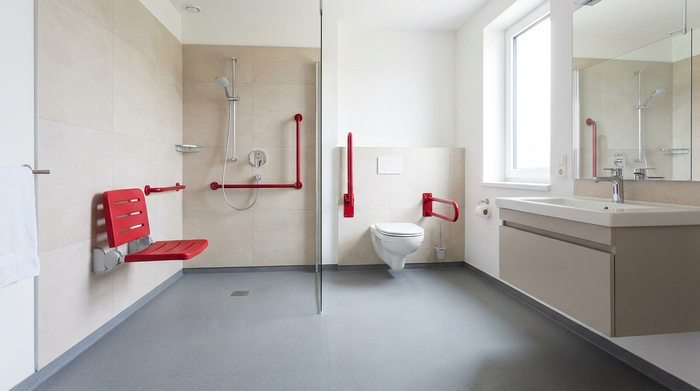 Ein modernes, barrierefreies Badezimmer mit einer ebenerdigen Dusche. Helle Fliesen an den Wänden, rutschfester matter Boden. Rote, gut sichtbare Haltegriffe an der Wand neben der Toilette und in der Dusche. Ein Duschsitz ist an der Wand montiert. Realistisch, sauber, sicher.