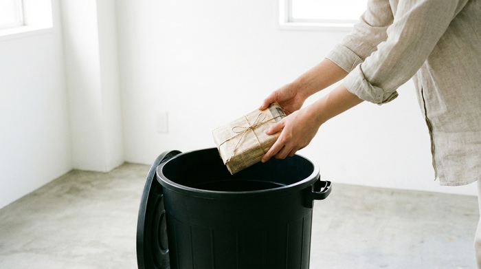 Eine Person wirft vorsichtig eine in neutrales Zeitungspapier eingewickelte kleine Schachtel in einen gewöhnlichen schwarzen Hausmülleimer. Sauberer, heller Hintergrund ohne Text.