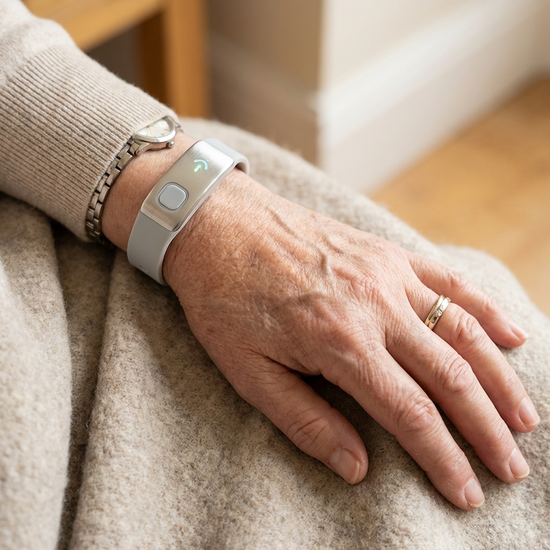 Ein moderner, unauffälliger Hausnotruf-Knopf am Handgelenk eines Senioren. Die Hand ruht entspannt auf einer weichen Decke. Helle, freundliche Farben.