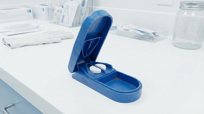 Ein spezieller, blauer Tablettenteiler aus Kunststoff liegt auf einer sauberen weißen Arbeitsfläche. Eine weiße Tablette ist perfekt in zwei Hälften geteilt. Medizinisch saubere Umgebung.