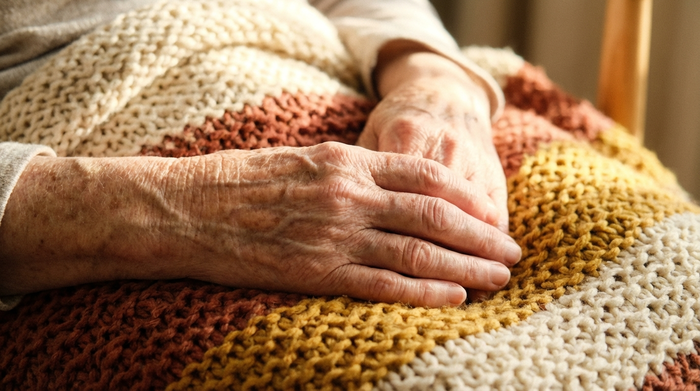 Eine Nahaufnahme von sanften, faltigen Händen eines Senioren, die ruhig auf einer weichen Kuscheldecke ruhen. Friedliche und schmerzfreie Stimmung im warmen Sonnenlicht.