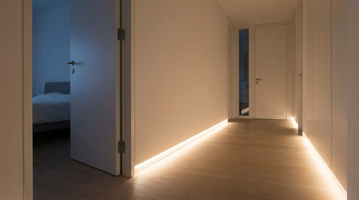 Eine sanfte, blendfreie LED-Beleuchtung entlang der Fußleisten in einem modernen, sauberen Flur bei Nacht. Ein sicherer Weg vom Schlafzimmer zum Bad wird ausgeleuchtet. Realistische Architekturfotografie.