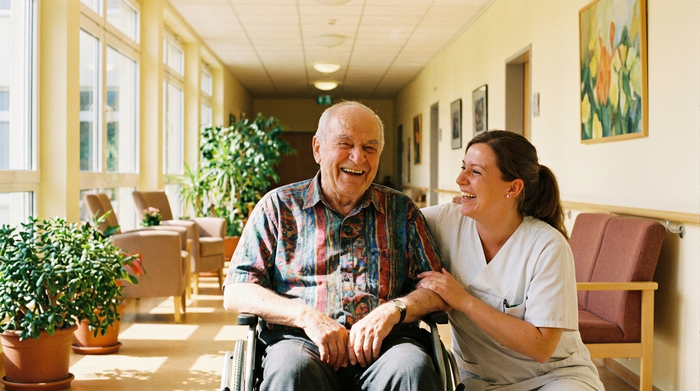 Ein fröhlicher älterer Herr im Rollstuhl lacht gemeinsam mit einer sympathischen Pflegekraft in einem sonnendurchfluteten Flur. Beide wirken entspannt und sehr vertraut miteinander.