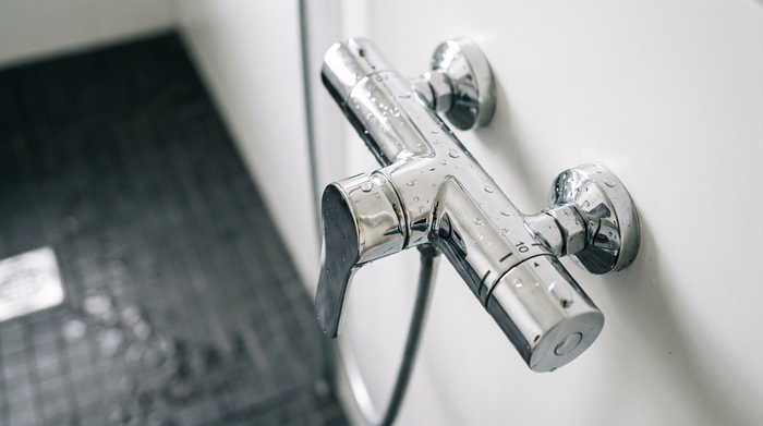 Nahaufnahme einer modernen, verchromten Thermostat-Duscharmatur mit einem langen Einhebelmischer an einer weißen Wand. Im Hintergrund sind unscharf rutschfeste, anthrazitfarbene Bodenfliesen einer bodengleichen Dusche zu erkennen.