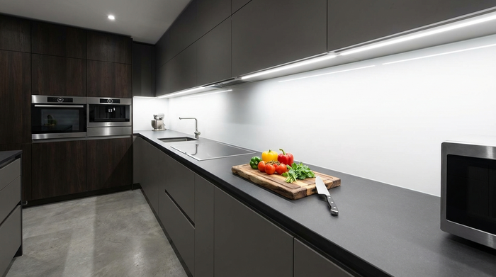 Eine aufgeräumte, moderne Küchenzeile. Unter den Hängeschränken sind durchgehende LED-Leisten angebracht, die die matte Arbeitsplatte extrem hell und völlig schattenfrei beleuchten. Frisches Gemüse und ein Messer liegen auf einem Holzschneidebrett.