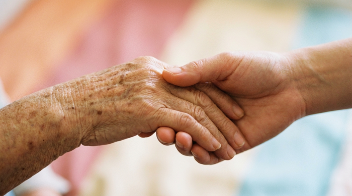 Nahaufnahme von zwei Händen, die sich sanft und tröstend berühren. Eine ältere, von Lebenserfahrung gezeichnete Hand wird von einer jüngeren Hand gehalten. Warmer, weicher Hintergrund in hellen Pastelltönen. Die Szene strahlt tiefe Geborgenheit und Fürsorge aus.